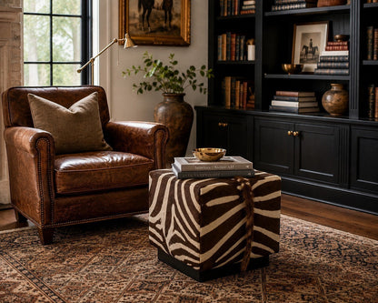 Pillowfina handcrafted vintage zebra hide cube ottoman styled in a luxury library with a brown leather armchair.